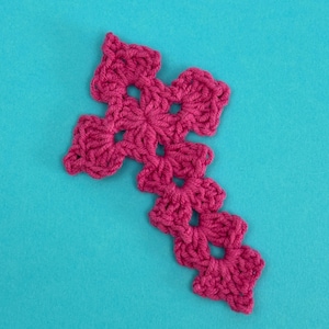 May include: A pink crocheted cross with a decorative design. The cross is made with a single crochet stitch and has a delicate, lacy appearance.