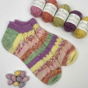 May include: A pair of hand-knitted ankle socks featuring a variegated pattern of pastel yellow, light green, soft pink, and lavender stripes. Five small balls of Scheepjes Metropolis yarn in coordinating colours and several speckled eggs are arranged nearby.