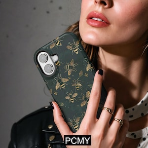 Puede incluir: Funda de teléfono verde oscuro con un patrón de abejas y hojas doradas. La funda tiene un recorte para la cámara y el texto "PCMY" en la parte inferior. La funda la sostiene una persona que lleva anillos y una chaqueta de cuero negra.