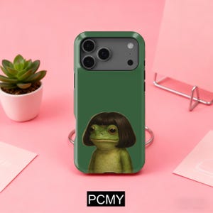 May include: A green phone case featuring a cartoon frog with a dark brown bob haircut. The frog has a neutral expression. The case has a dark green border and the text "PCMY" at the bottom.