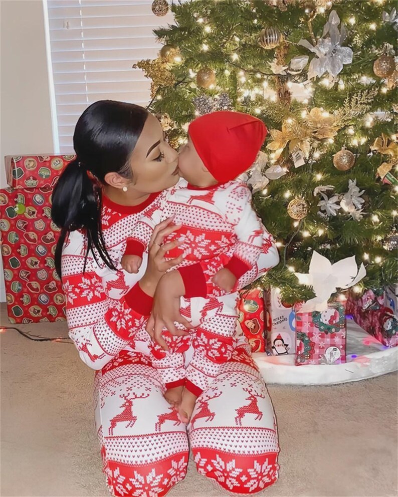 Puede incluir: Una mujer y un beb&eacute; con pijamas navide&ntilde;os rojos y blancos a juego, bes&aacute;ndose frente a un &aacute;rbol de Navidad decorado. Los pijamas presentan un estampado de renos y copos de nieve. El beb&eacute; lleva un gorro rojo.