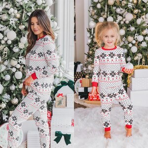 Puede incluir: Conjunto de pijamas navide&ntilde;os blancos a juego con un estampado de copos de nieve. Las camisetas y pantalones de manga larga tienen ribetes rojos en los pu&ntilde;os y el cuello. Los pijamas los llevan una mujer y una ni&ntilde;a peque&ntilde;a.