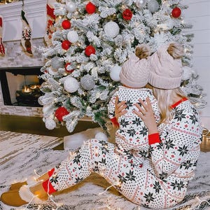 Puede incluir: Una madre y un ni&ntilde;o con pijamas de copos de nieve a juego y gorros de punto con pompones se sientan frente a un &aacute;rbol de Navidad decorado. El &aacute;rbol est&aacute; adornado con adornos rojos, blancos y plateados. Una chimenea y calcetines son visibles en el fondo.