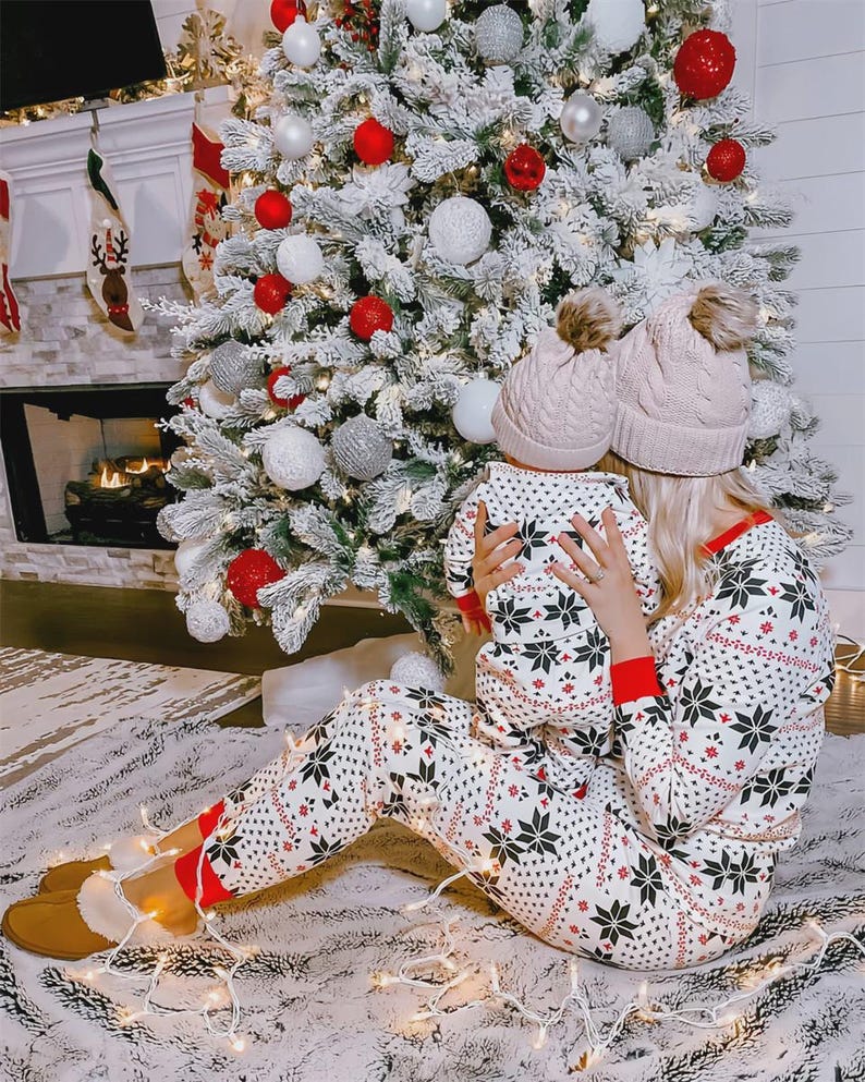 Puede incluir: Una madre y un ni&ntilde;o con pijamas de copos de nieve a juego y gorros de punto con pompones se sientan frente a un &aacute;rbol de Navidad decorado. El &aacute;rbol est&aacute; adornado con adornos rojos, blancos y plateados. Una chimenea y calcetines son visibles en el fondo.