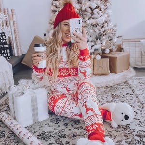 Puede incluir: Una persona con gorro rojo y un pijama a juego rojo y blanco con motivos navide&ntilde;os y un estampado de renos. La persona sostiene una taza de caf&eacute; y un tel&eacute;fono. Un &aacute;rbol de Navidad y regalos envueltos en el fondo.