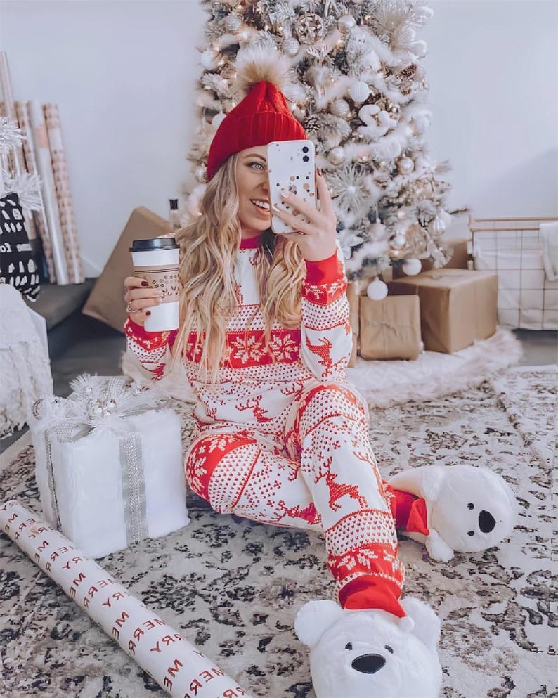 Puede incluir: Una persona con gorro rojo y un pijama a juego rojo y blanco con motivos navide&ntilde;os y un estampado de renos. La persona sostiene una taza de caf&eacute; y un tel&eacute;fono. Un &aacute;rbol de Navidad y regalos envueltos en el fondo.
