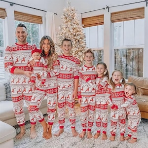 Puede incluir: Un grupo de personas con pijamas navide&ntilde;os rojos y blancos a juego. Los pijamas presentan un dise&ntilde;o de renos y copos de nieve. La familia est&aacute; de pie frente a un &aacute;rbol de Navidad.
