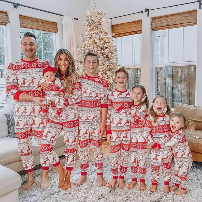 Puede incluir: Un grupo de personas con pijamas navide&ntilde;os rojos y blancos a juego. Los pijamas presentan un dise&ntilde;o de renos y copos de nieve. La familia est&aacute; de pie frente a un &aacute;rbol de Navidad.