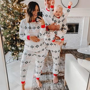 Puede incluir: Una familia con pijamas blancos a juego con un estampado de copos de nieve y ribetes rojos. Los pijamas incluyen tops de manga larga y pantalones. La familia sonr&iacute;e y posa para una foto frente a un &aacute;rbol de Navidad y una chimenea.