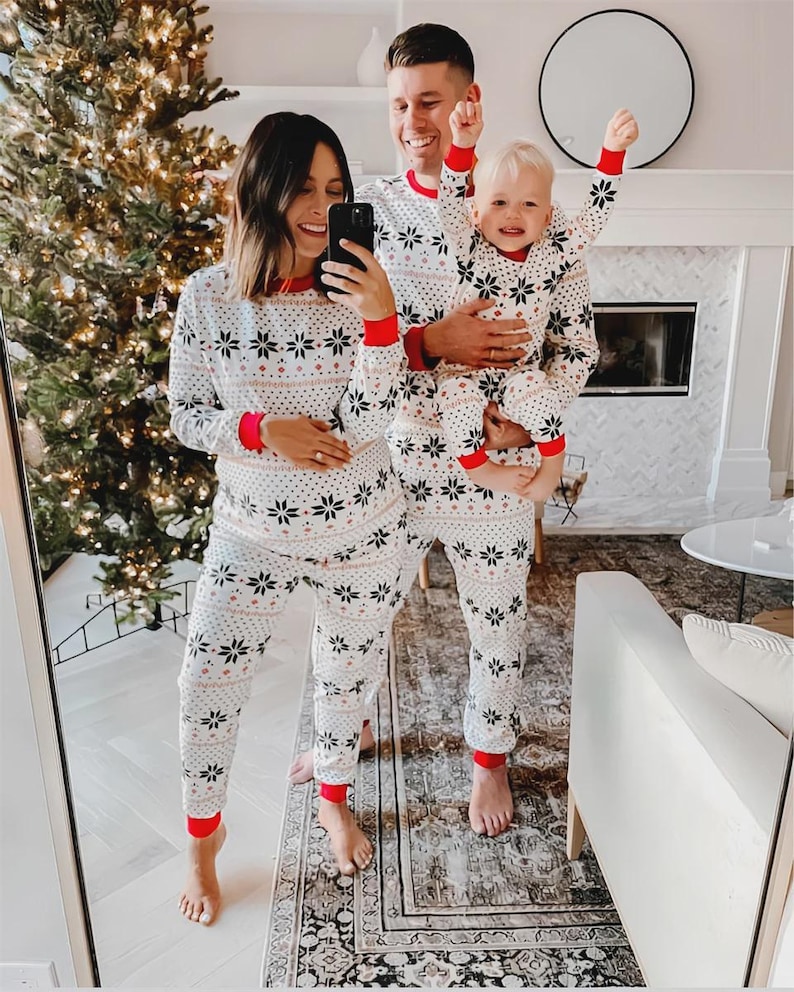 Puede incluir: Una familia con pijamas blancos a juego con un estampado de copos de nieve y ribetes rojos. Los pijamas incluyen tops de manga larga y pantalones. La familia sonr&iacute;e y posa para una foto frente a un &aacute;rbol de Navidad y una chimenea.