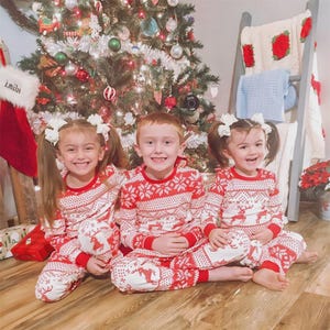 Puede incluir: Tres ni&ntilde;os con pijamas navide&ntilde;os rojos y blancos a juego sentados frente a un &aacute;rbol de Navidad decorado. Los pijamas presentan un estampado de copos de nieve y renos. Una media roja con el nombre "Emilit" cuelga cerca.