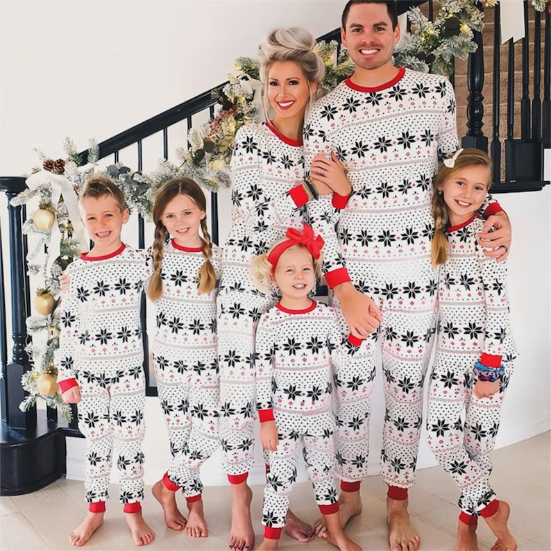 Puede incluir: Una familia de siete personas con pijamas a juego blancos y negros con estampado de copos de nieve y ribetes rojos. Los pijamas incluyen tops de manga larga y pantalones. La familia est&aacute; de pie frente a una escalera decorada con guirnaldas navide&ntilde;as.