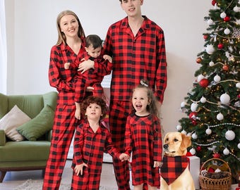 May include: A family wearing matching red and black plaid pajamas. The group includes two adults, three children, and a dog wearing a matching bandana. A Christmas tree with ornaments is in the background.