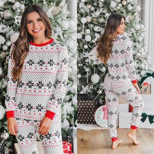 Puede incluir: Un conjunto de pijama a juego con un fondo blanco y un patr&oacute;n repetido de copos de nieve negros y detalles rojos. La parte superior de manga larga y los pantalones tienen ribetes rojos. La modelo posa frente a un &aacute;rbol de Navidad.