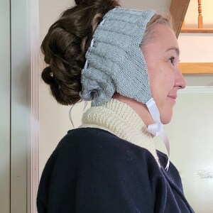May include: A gray knitted headband with a white ribbon tied at the back. The headband is worn over a woman's hair, which is styled in a bun.