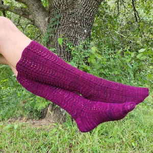 May include: A pair of long, purple knitted socks with a textured pattern. The socks are worn by a person sitting on the ground with their legs crossed.