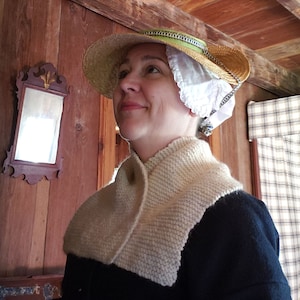 May include: A woman wearing a white knitted shawl and a straw hat with a white lace trim. She is standing in a room with wooden walls and a plaid curtain.