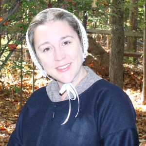 Puede incluir: Una mujer con un gorro de crochet blanco y una chaqueta azul oscuro con una chal marr&oacute;n. Est&aacute; de pie en un &aacute;rea boscosa con hojas de oto&ntilde;o en el suelo.