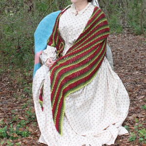 May include: A woman wearing a white dress with a floral pattern is sitting in a chair. She is wearing a green, red, and yellow striped shawl.