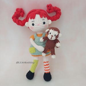 May include: A handmade crochet doll with red pigtails, wearing a green dress and holding a brown monkey. The doll has a white face, orange shirt, and striped socks. The doll's legs are green and orange. The doll's feet are black. The doll is a handmade item.