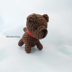 May include: A handmade brown crochet dog with a red collar. The amigurumi dog has black eyes and a black nose. The text "@leahknitting" is visible in the lower left corner. The dog is standing on a light blue surface.
