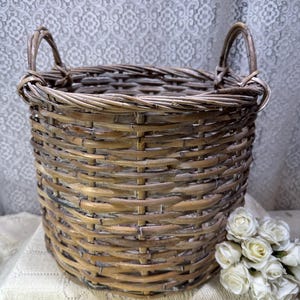 May include: A round, woven wicker basket with two arched handles. The basket is a natural tan color and has a textured surface. A small bouquet of white roses sits next to the basket.