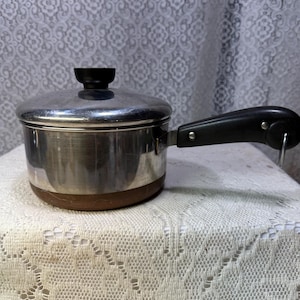 May include: A stainless steel saucepan with a lid and a black handle. The pot has a copper-colored bottom and a black knob on the lid. The handle is attached with rivets. The pot is sitting on a white lace tablecloth.