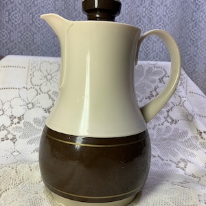 May include: A vintage coffee carafe with a cream-colored upper section and a dark brown lower section. The carafe has a matching brown lid and handle. A thin gold-colored band accents the brown section. The carafe is likely made of ceramic or a similar material.