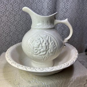 May include: A cream-colored ceramic pitcher and basin set featuring a speckled finish. The pitcher has a prominent embossed rose design on the front, while the basin displays a decorative scalloped edge along the rim, creating a cohesive set.