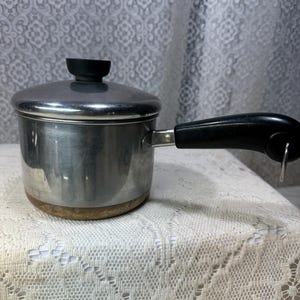 May include: A small, stainless steel saucepan with a lid and a black handle. The pot has a silver finish and a black knob on the lid. The handle is attached to the side of the pot. The pot is sitting on a white lace tablecloth.