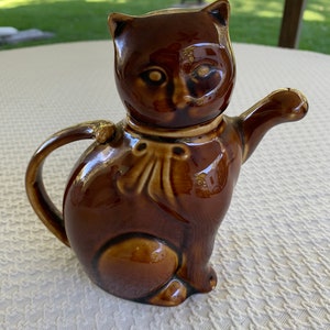 May include: A brown ceramic cat teapot with a raised tail and a brown ribbon around its neck. The teapot has a spout on the side and a lid on top.