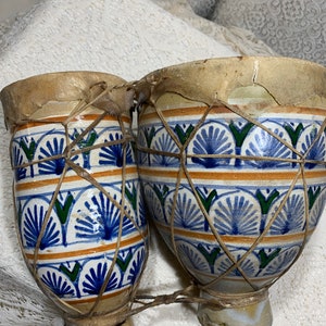 May include: A pair of ceramic bongo drums with a decorative blue, green, and orange floral pattern. The drums have tan leather drumheads and are connected by a woven cord. The drums are set against a white background.