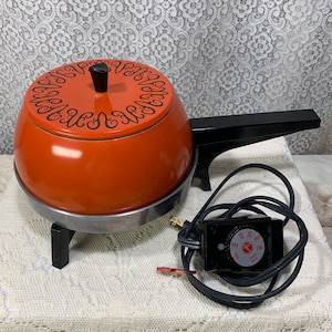 May include: Orange electric fondue pot with a black handle and a black temperature control knob. The pot has a decorative design around the top and sits on three black legs.