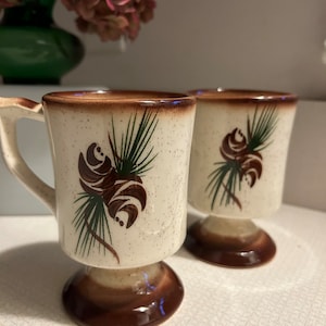 May include: Two ceramic coffee mugs with brown and green pine cone designs. The mugs have a brown speckled glaze and a brown pedestal base.