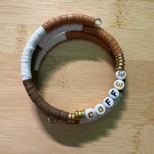 May include: A handmade bracelet featuring brown, white, and gold beads. The bracelet spells out the word "COFFEE" with white letter beads. The bracelet is made of flat, round beads and is designed to be worn around the wrist.