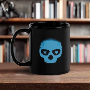 May include: A black ceramic mug featuring a blue skull design. The skull has an X for one eye and a heart for the other. The mug is sitting on a wooden surface with a bookshelf in the background.