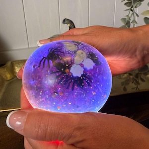 Peut inclure: Un presse-papier sphérique en verre avec un effet lumineux violet et bleu. À l'intérieur de l'orbe transparent se trouvent des silhouettes d'araignées sombres, de petites formes circulaires blanches et des paillettes multicolores éparpillées.