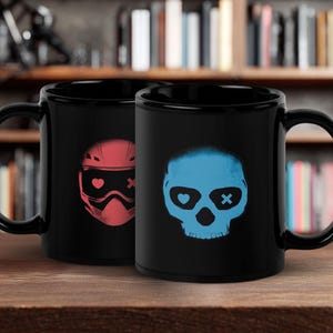 May include: Two black ceramic mugs with unique designs. One mug features a red helmet graphic with a heart and an X, while the other displays a blue skull with a heart and an X. Both mugs have a glossy finish.