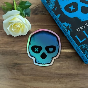 May include: A holographic skull sticker with a blue and purple gradient. The skull has an X for one eye and a heart for the other. A white rose and a book titled "Lights Out" are also in the image.