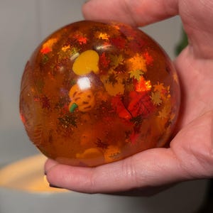 May include: A spherical resin paperweight with a translucent amber tint, containing embedded miniature orange pumpkin shapes and small, multicolored autumn leaf confetti, featuring a smooth, glossy surface and reflective glitter accents.