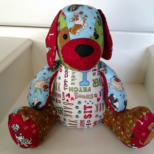 May include: A stuffed dog toy with a patchwork design. The dog has a red nose, ears, and paws. The body is covered in fabric with dog-themed patterns and text, including paw prints, bones, and the words "Bark" and "Fetch".