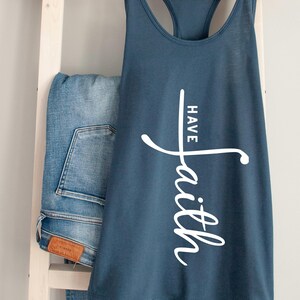 Könnte beinhalten: Ein marineblaues Tanktop mit dem Schriftzug "Have Faith" in weißer Schrift. Das Tanktop hängt an einem Holzkleiderbügel, mit einer Jeans und einem braunen Hut im Hintergrund. Das Tanktop ist ein lässiger Stil.