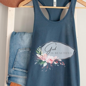 Könnte beinhalten: Ein tealfarbenes Tanktop mit floralem Design und dem Schriftzug "God is Beautiful". Das Tanktop hängt an einem Holzkleiderbügel. Ein brauner Hut und blaue Jeans sind ebenfalls im Bild zu sehen.