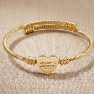 May include: Gold-colored bangle bracelet with a heart-shaped charm. The charm is engraved with the words "Consider the Wildflowers" and "Luke 12:27". The bracelet has a twisted cable design and decorative accents.