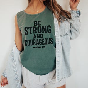 May include: A green tank top with the words "BE STRONG AND COURAGEOUS" in black block letters. The text "Joshua 1:9" is below the main text. A light blue denim jacket is worn over the tank top.