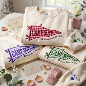 May include: Cream-colored sweatshirts with embroidered pennant designs reading "Camp Sophia" in various colors. The pennants feature gingham patterns and the text "Miami Beach, FL 2026" or "Miami Beach, FL 202". Matching wrist cuffs are also visible.