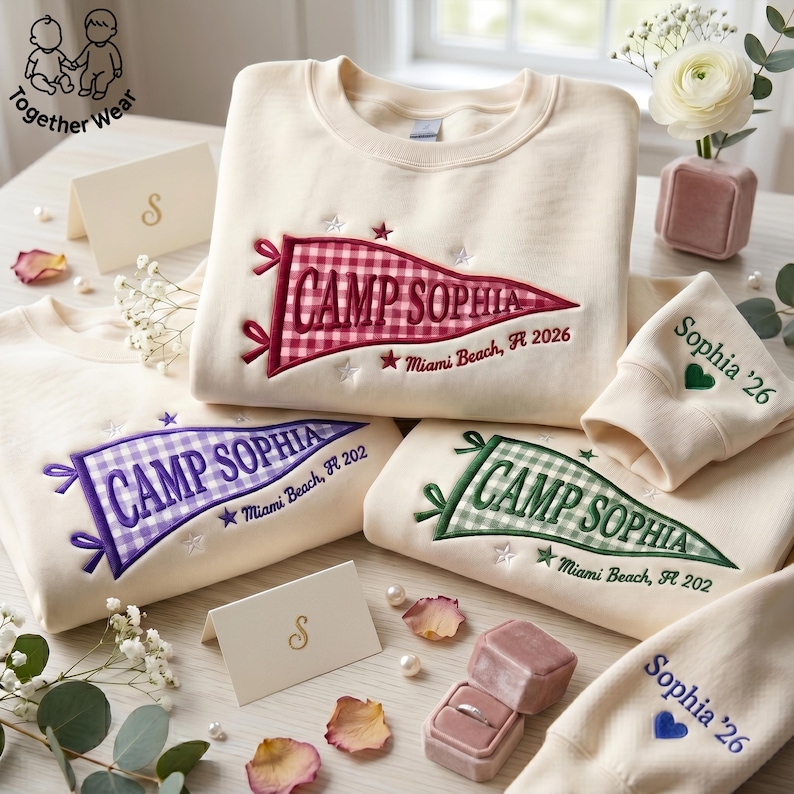 May include: Cream-colored sweatshirts with embroidered pennant designs reading "Camp Sophia" in various colors. The pennants feature gingham patterns and the text "Miami Beach, FL 2026" or "Miami Beach, FL 202". Matching wrist cuffs are also visible.