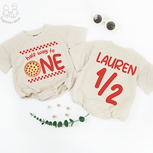 May include: Two beige baby rompers. One says "half way to ONE" with a pizza graphic. The other says "LAUREN 1/2" in red. Sunglasses and decorative elements are also present.
