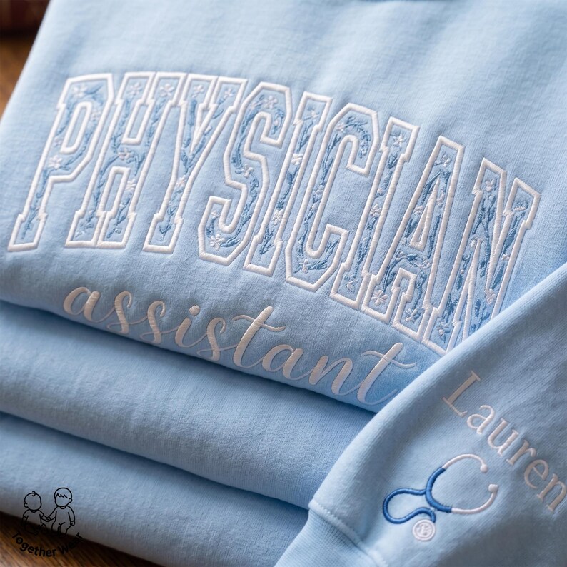 May include: Light blue sweatshirts with white embroidered text. The top sweatshirt reads "PHYSICIAN" in a decorative font, with "assistant" below. A smaller sweatshirt has "Lauren" and a stethoscope design. The sweatshirts are folded.