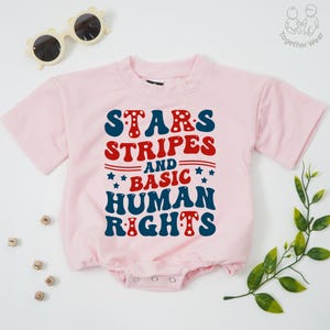 May include: A light pink baby bodysuit with the text "STARS STRIPES AND BASIC HUMAN RIGHTS" in red and blue. The bodysuit has short sleeves and a snap closure. Sunglasses are in the top left corner.